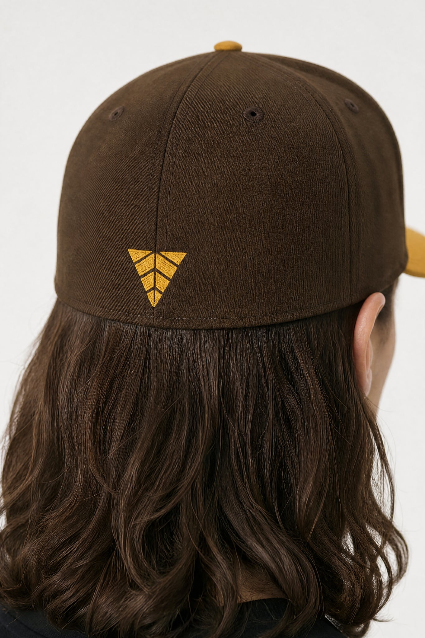 Brown cap with a yellow logo worn by a person with long hair on a white background