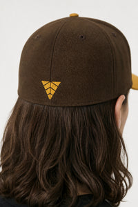 Brown cap with a yellow logo worn by a person with long hair on a white background