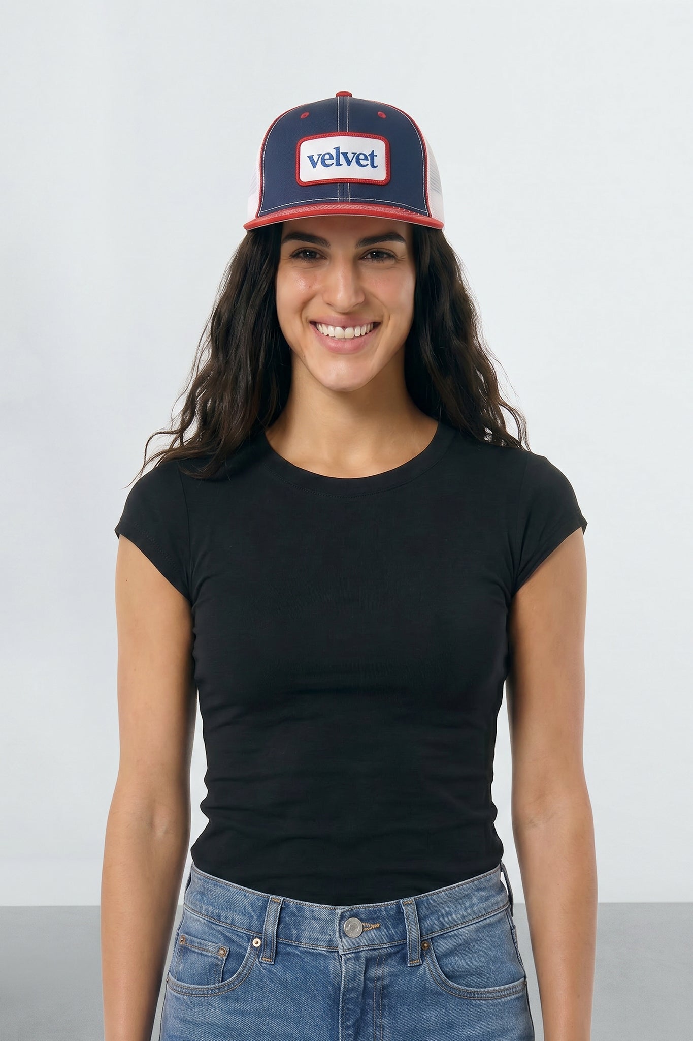 Woman wearing a black t-shirt and blue jeans with a navy and red cap on a white background