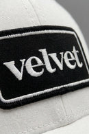 White cap with a black patch displaying the word 'velvet'.